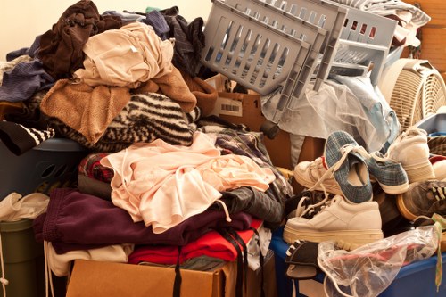 A large collection of mixed household items and clothing arranged in a cluttered pile, including neatly folded and crumpled garments in various fabrics and colours. On the left side, there are soft textiles such as sweaters and t-shirts, with some pieces draped over the pile. A grey plastic laundry basket is positioned towards the top right, filled with additional textiles or discarded items, with some clothes spilling over its edges. Nearby, a white sneaker and a pair of beige shoes are visible among the clutter, along with a transparent plastic bag containing more waste. The items are situated on a surface that appears to be an indoor floor or a storage area, with a warm light illuminating the scene. This image relates to rubbish removal services offered by Balham House Clearance, situated near postcode SW12, London, highlighting the need for professional clearance of household clutter and waste.