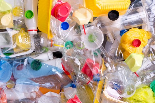 A close-up view of a large pile of discarded plastic bottles and containers, primarily transparent with some yellow, red, green, and blue caps, mixed together chaotically on a flat surface. The bottles vary in size and shape, with some crushed or deformed and others retaining their original form. Many have visible labels, some partially torn or faded. The image captures the texture of the crinkled plastic, highlighting the glossy, reflective surfaces under natural light. In the background, there is no distinct environmental context, focusing solely on the heap of waste plastic. This visually accurate depiction relates to rubbish removal services like those offered by Balham House Clearance, emphasizing the importance of proper disposal and recycling of plastic waste in the local area, such as in the Balham region of London postcode SW12.