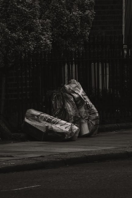 A collection of scattered domestic rubbish and discarded items is shown piled on gravel ground next to a low stone wall. The debris includes several black plastic garbage bags, crumpled white and clea